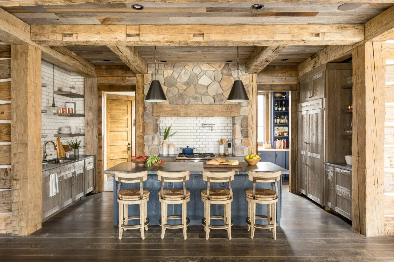 Weathered dark wood cabinets and a blue island sit beneath large wood beams, with a stone hood surround and white tile.