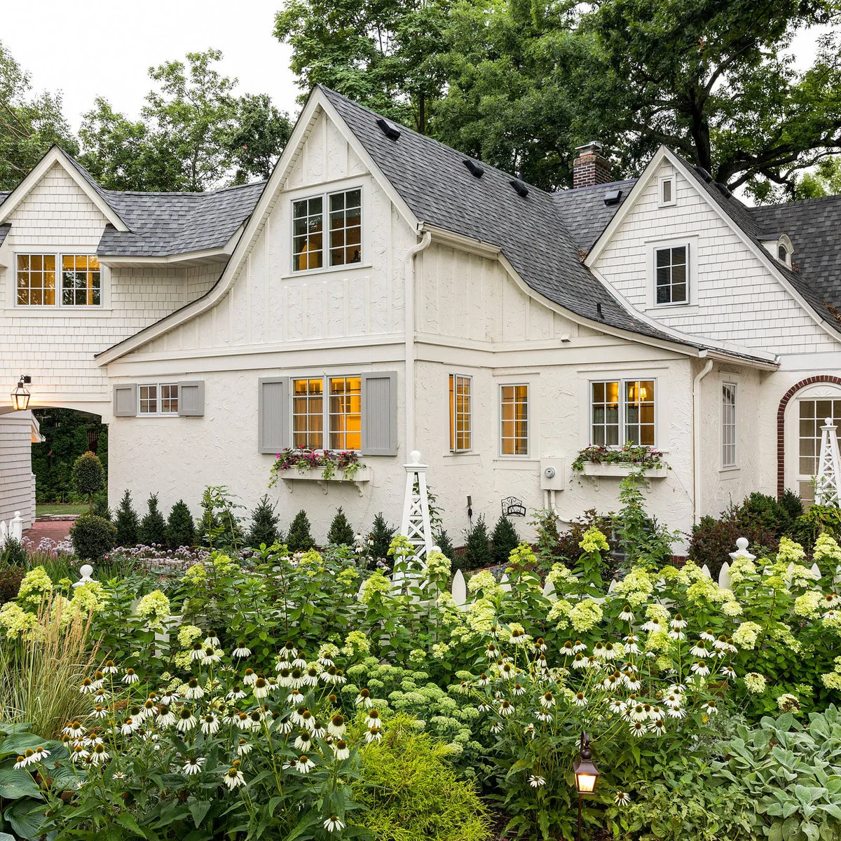 White window boxes and trellises rise above a dense cottage border of white flowers, broad leaves and silvery foliage.