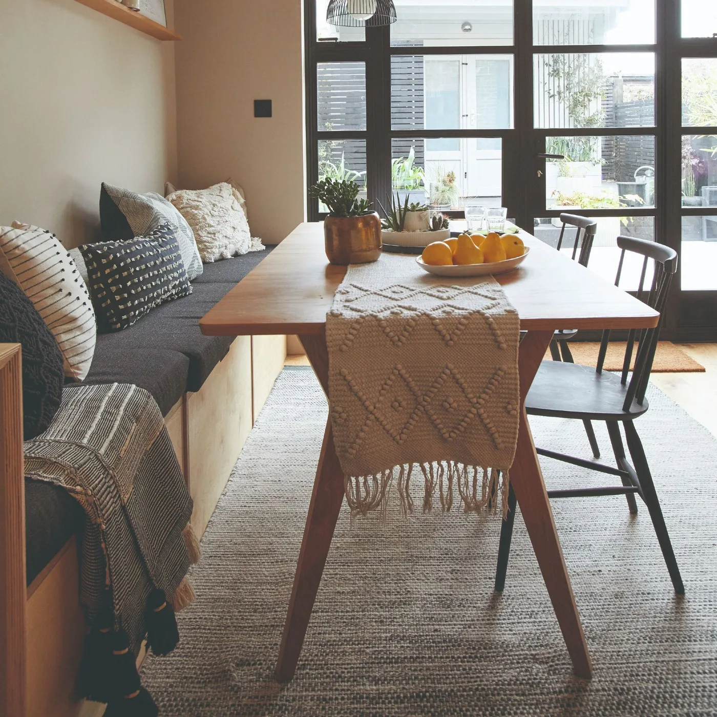 Dining room with a wooden table, dark spindle-back chairs, charcoal banquette, patterned cushions and black-framed glazed doors.