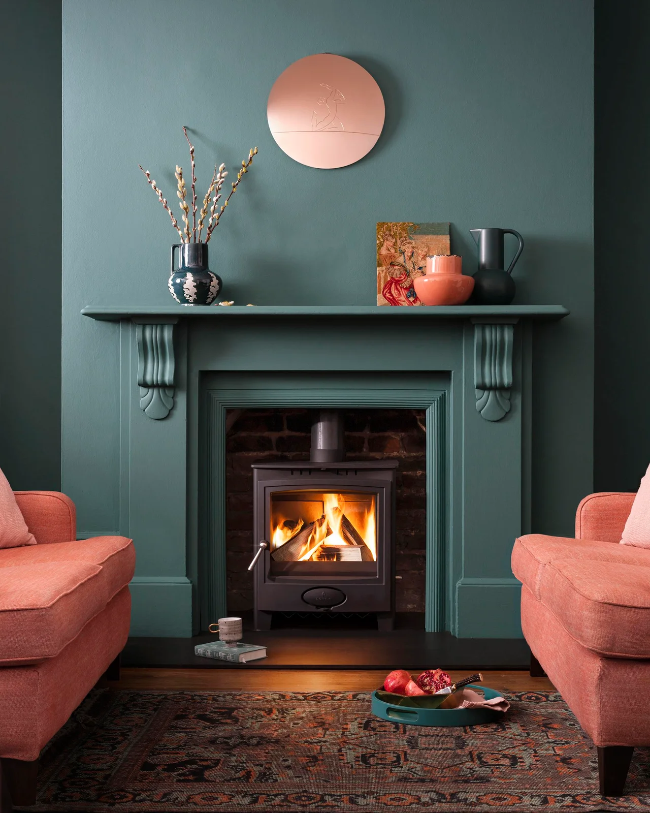 A deep teal mantel and wall frame a dark stove between salmon-colored seats, with exposed brick inside the opening.