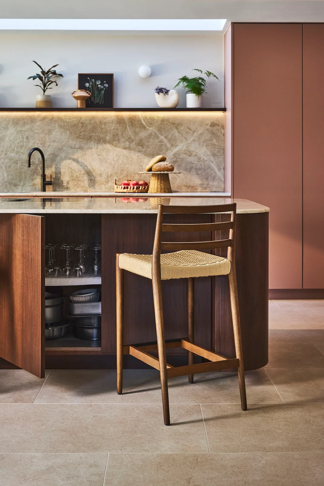 An open walnut island cupboard holds glasses and cookware beside a woven stool, with pink cabinets and a veined backsplash behind.