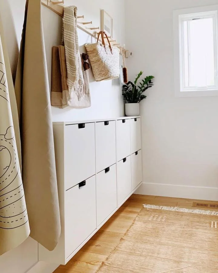 White wall-mounted shoe cabinets beneath a long wood peg rail holding bags and textiles beside an entryway window.