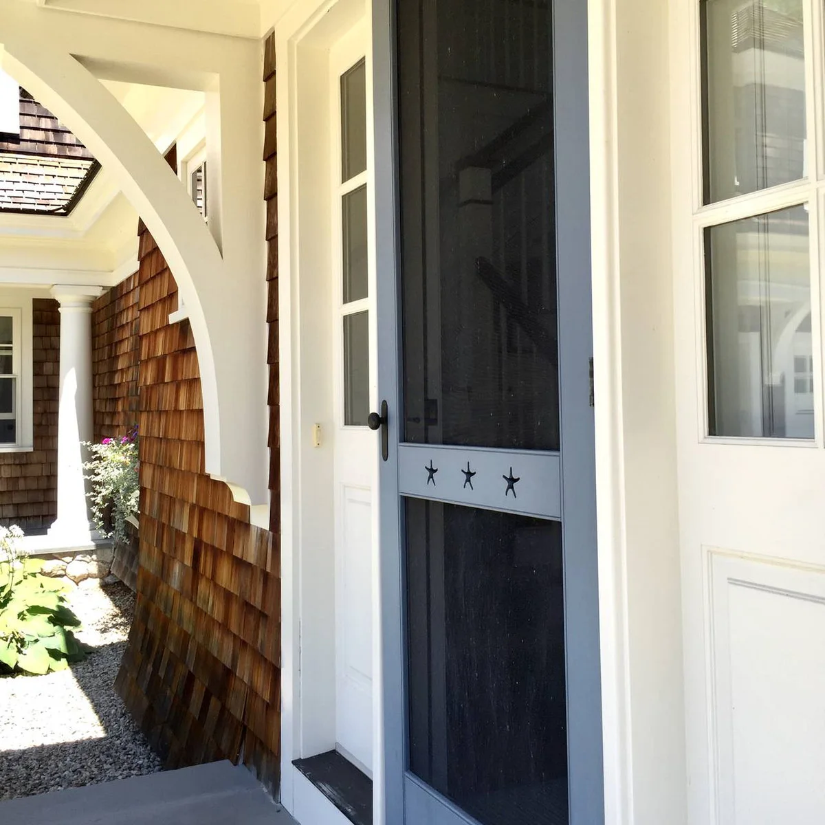 A dark blue screen door with three star-shaped details stands beside brown shingle siding, white trim and a curved white porch bracket.