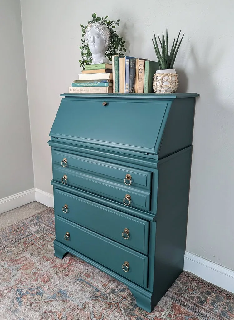 Closed blue-green secretary desk with gold-toned ring pulls and books and plants arranged on top.