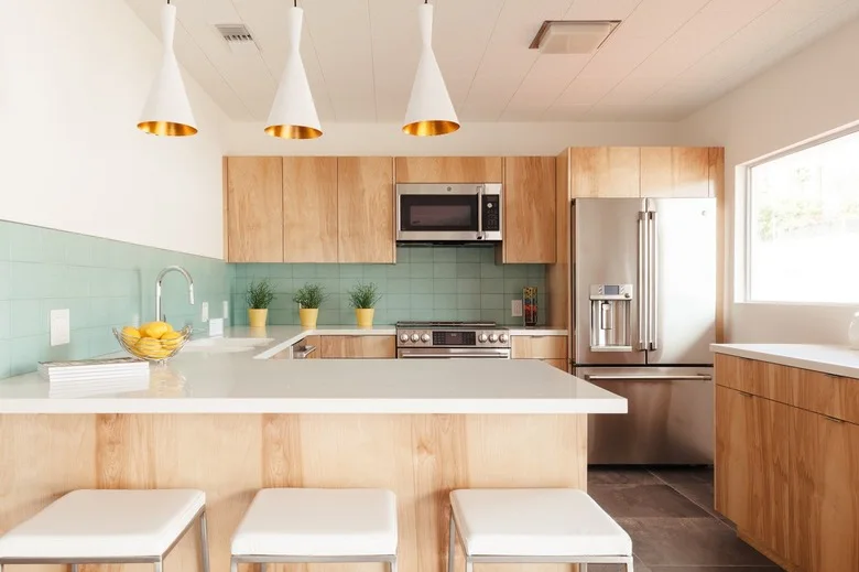 A pale wood kitchen has a mint-green rectangular backsplash, white counters, three white pendants with warm-colored interiors and a three-seat peninsula.