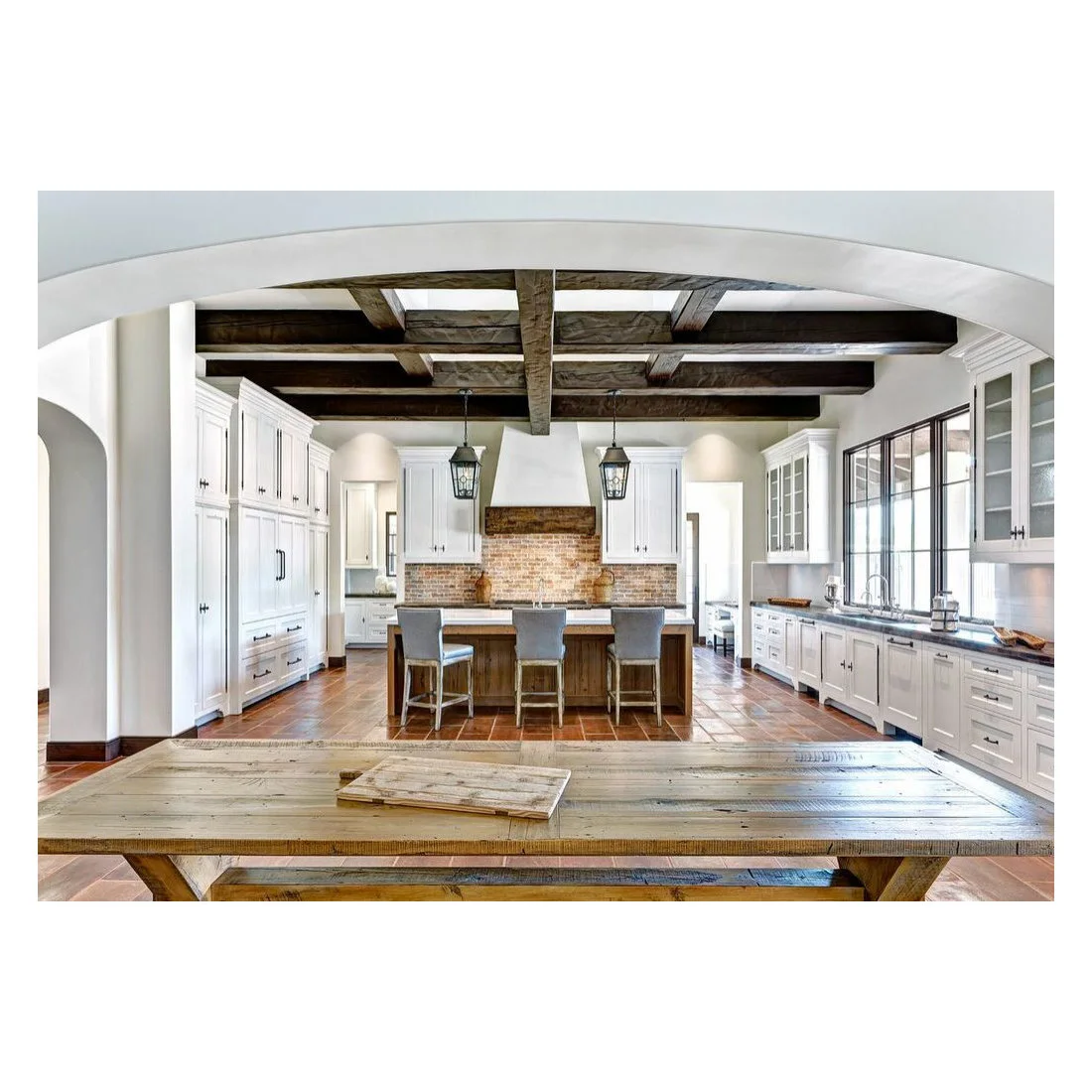 Dark ceiling beams run above white kitchen cabinets, a timber island and a broad timber table in the foreground.