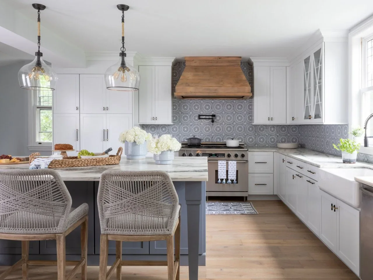 White kitchen cabinets frame a timber range hood, patterned gray backsplash and woven island stools.