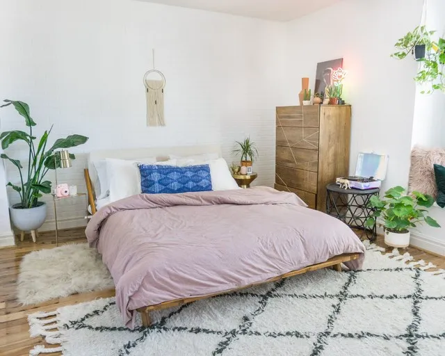 A low pale wood bed with a muted pink cover stands beside a tall wood chest, leafy plants and a small woven wall decoration against white walls
