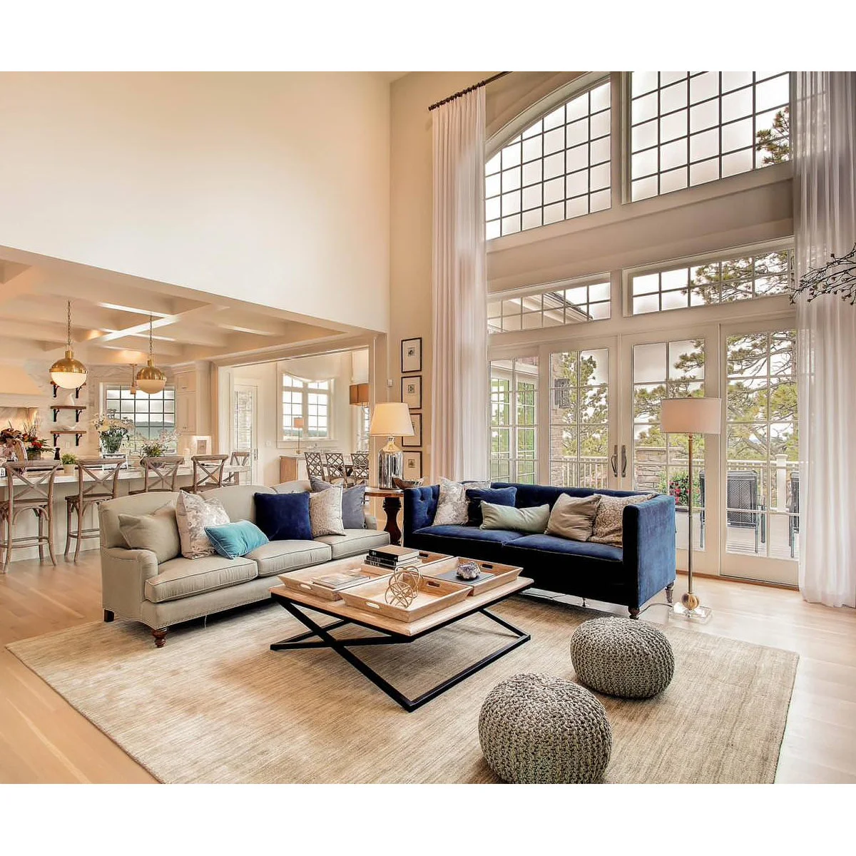A high living room with an arched upper window, long white curtains, blue and pale sofas and two knitted poufs.