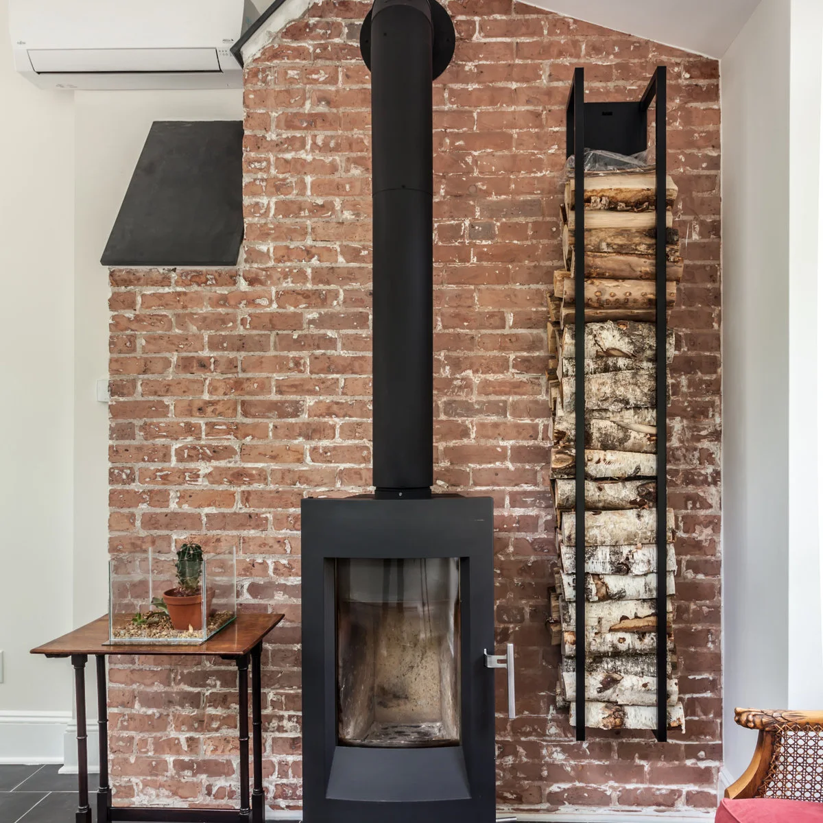 A tall black wood stove stands before exposed red brick, with a vertical log rack on one side and a small table on the other.