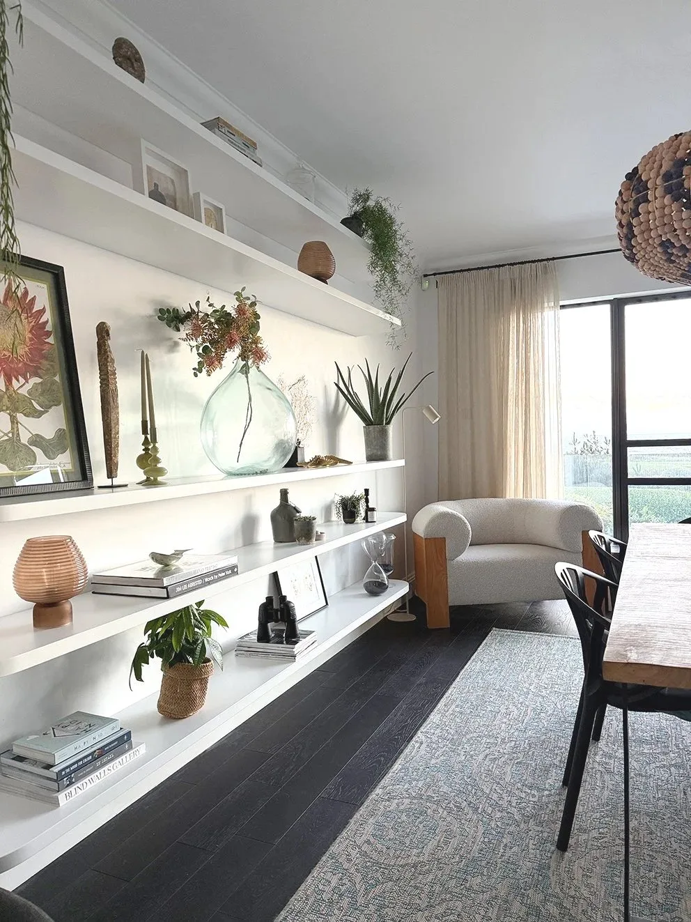 Long white shelves with rounded ends holding books, framed art and plants beside a wooden dining table and pale armchair