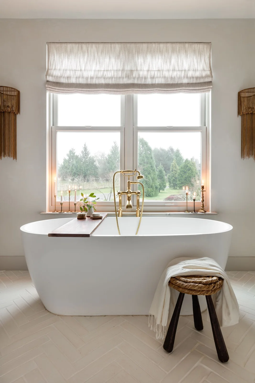 A plain white oval bath stands beneath a broad window beside a woven-seat wood stool, a wood bath tray, textured wall lights and a pale window shade