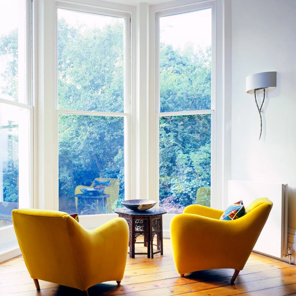 Two yellow armchairs angle toward a small dark side table inside a tall, white-framed bay window.