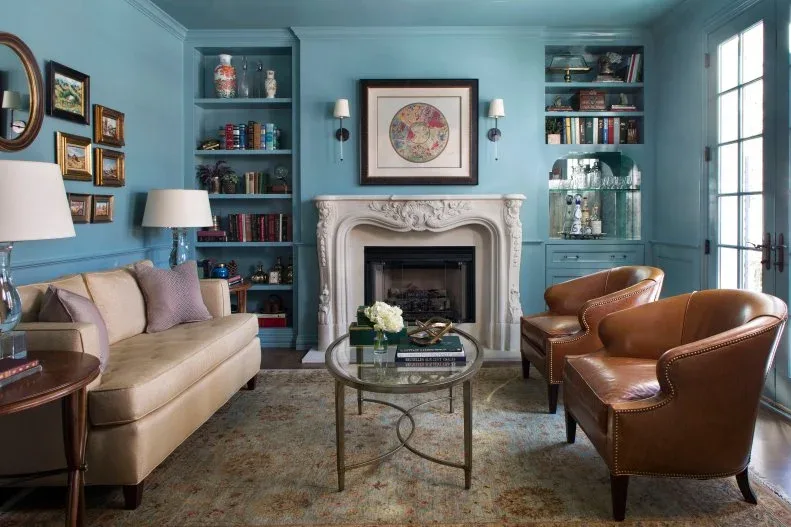 A carved pale limestone fireplace stands between blue built-in shelves beneath framed artwork.