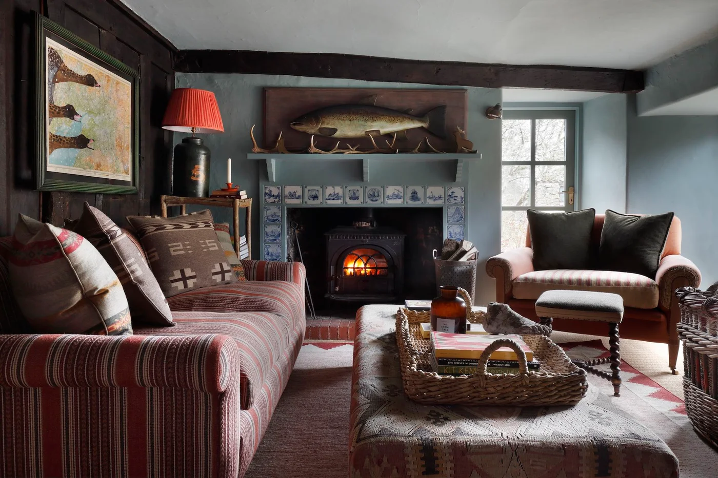Blue-and-white illustrated tiles border a stove opening beneath a painted mantel in a room with striped seating, dark beams and a red lamp.