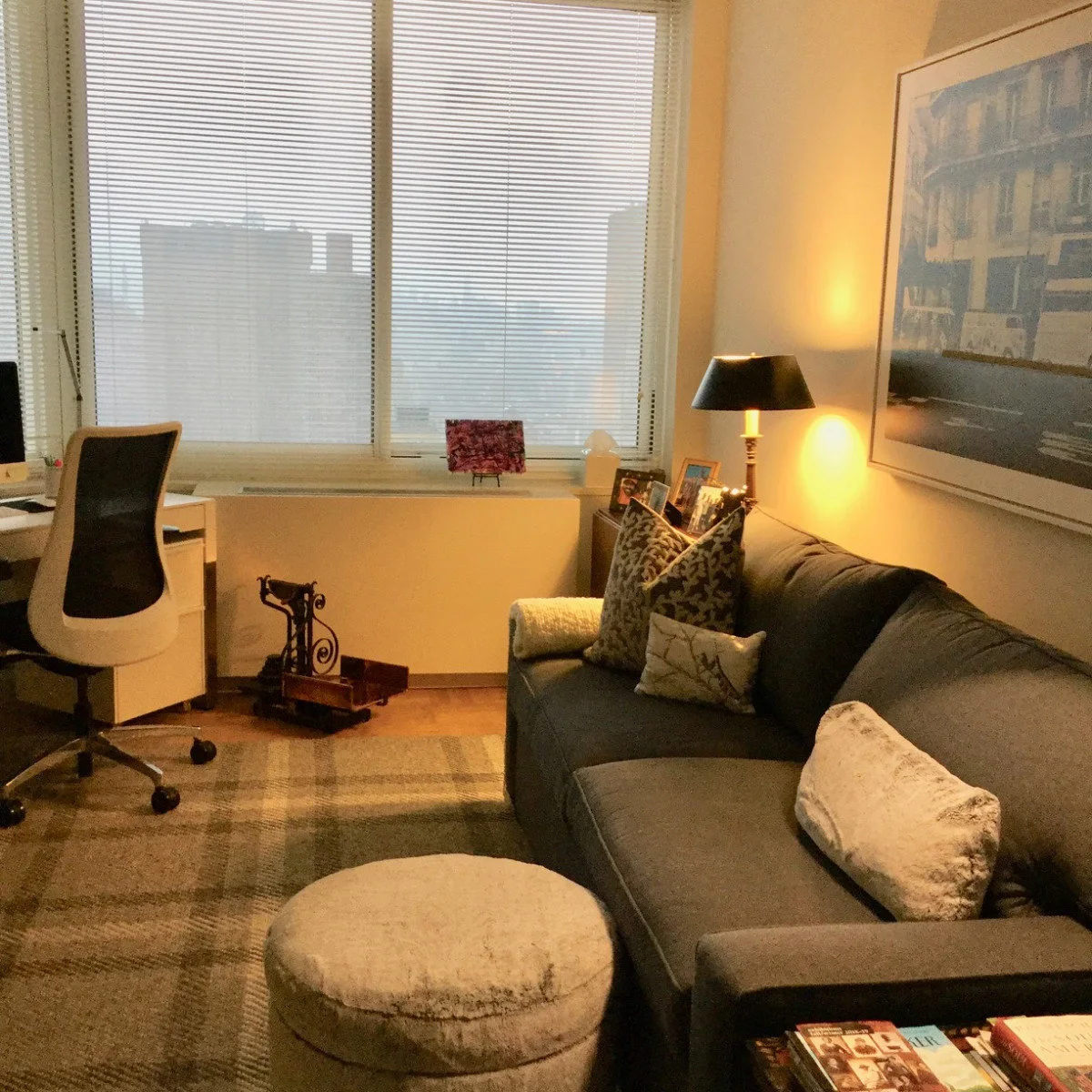 Dark gray sleeper sofa beside a desk, a round upholstered ottoman and a lit table lamp