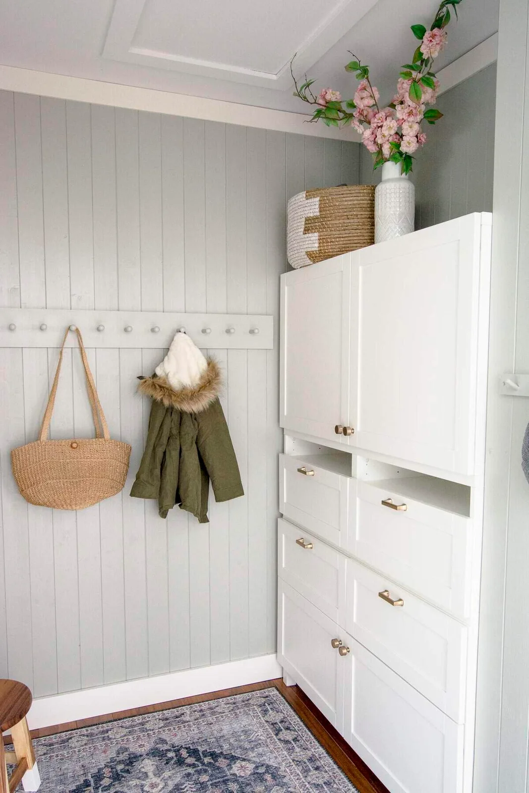 White mudroom cabinet with drawers, open slots and upper doors beside a peg rail on a pale gray paneled wall.