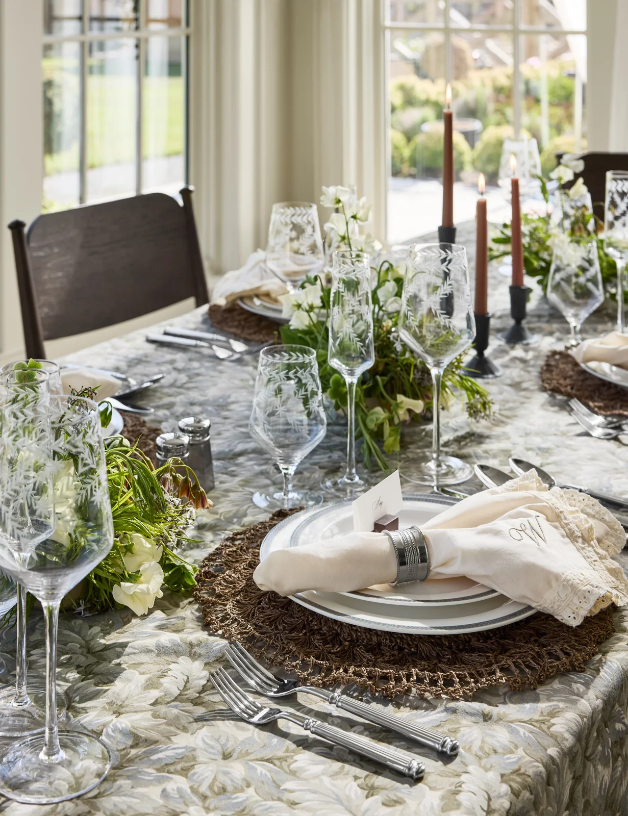 White plates and lace-edged napkins sit on woven mats above a muted green patterned cloth beside clear glassware