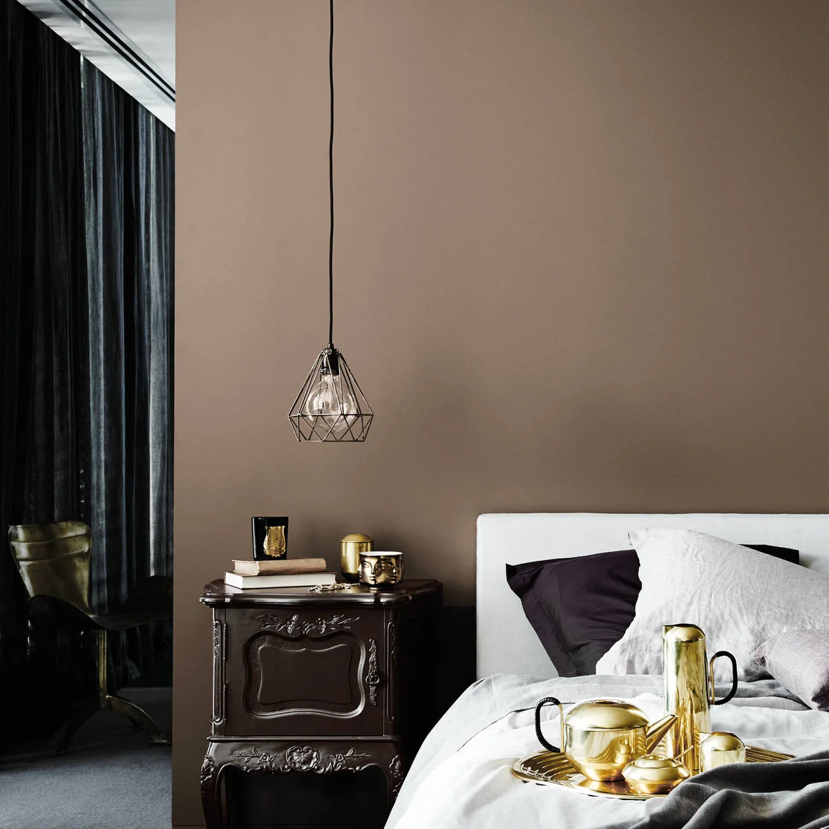 Muted brown bedroom wall with a dark carved bedside chest, white headboard and small gold-colored accessories.