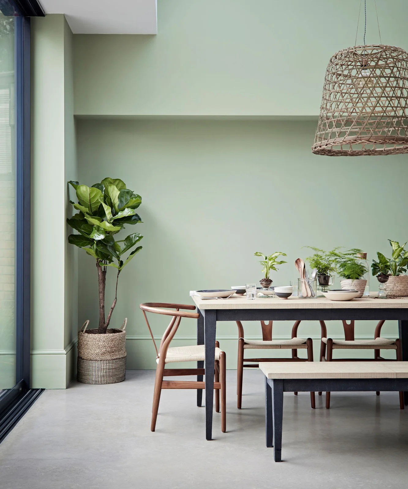Pale green walls surround a wood-topped table with dark legs, matching bench, wooden chairs and a woven-looking pendant