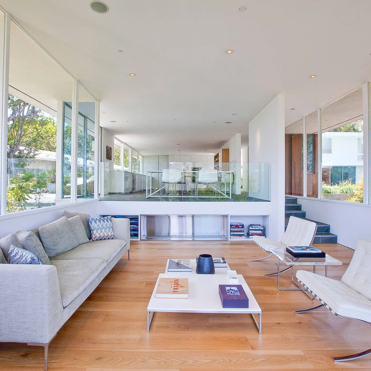 Elongated glazed interior with a gray sofa and white chairs below a raised glass-edged dining platform.
