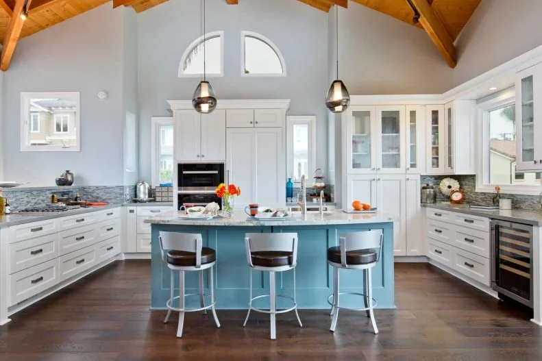 A blue paneled island with pale stools sits within white framed kitchen cupboards beneath a warm wood vaulted ceiling and two smaller hanging lights