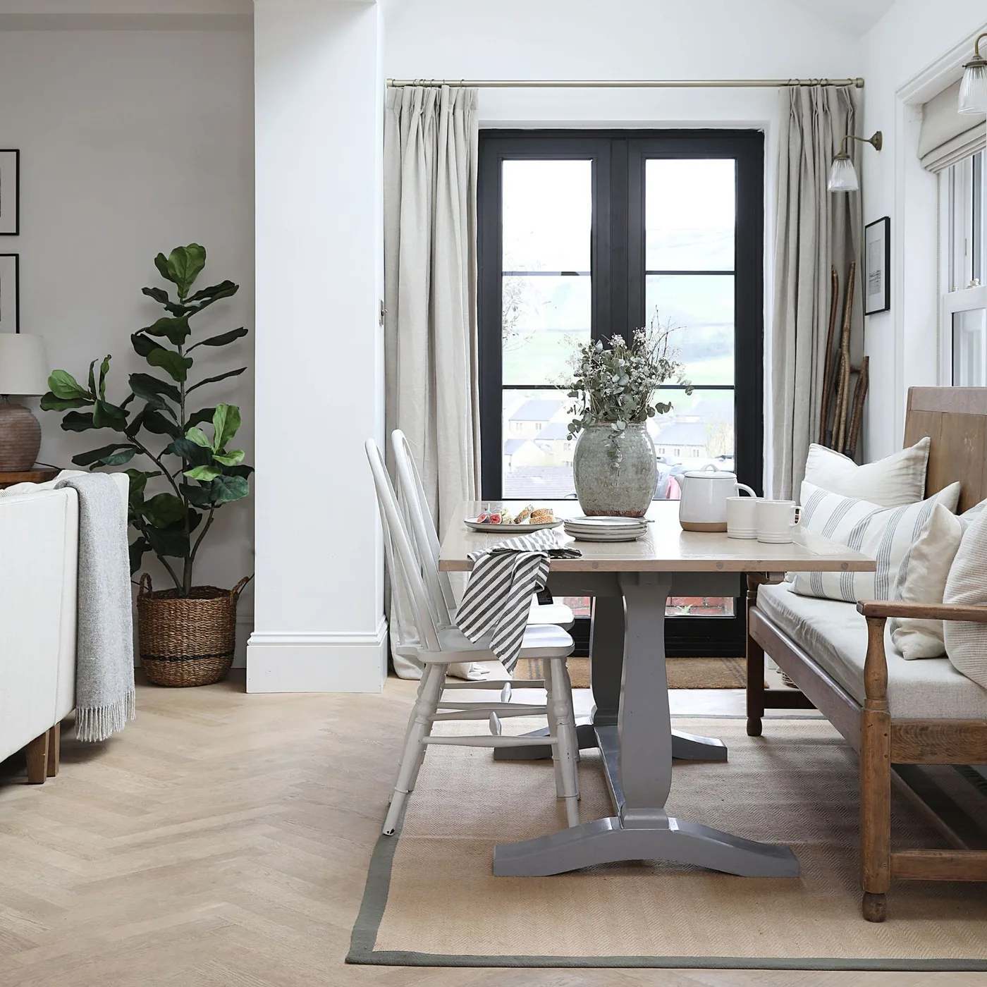 Open living and dining room with a large wooden table, white chairs, an upholstered wooden bench, pale rug and long curtains.