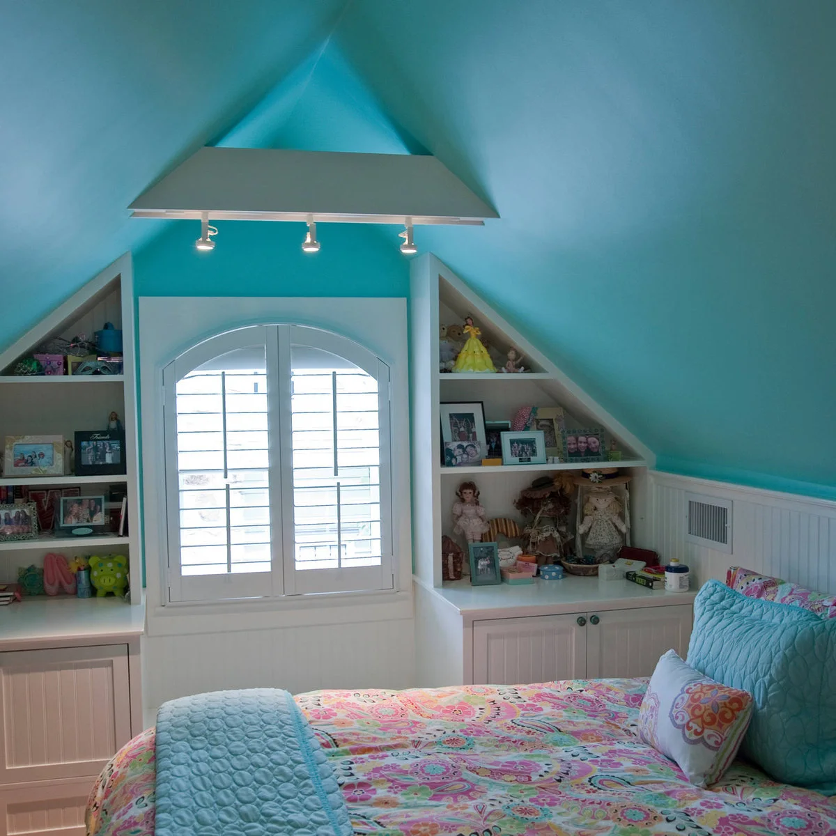 Turquoise attic bedroom with triangular shelving beside an arched window and a floral-covered bed.