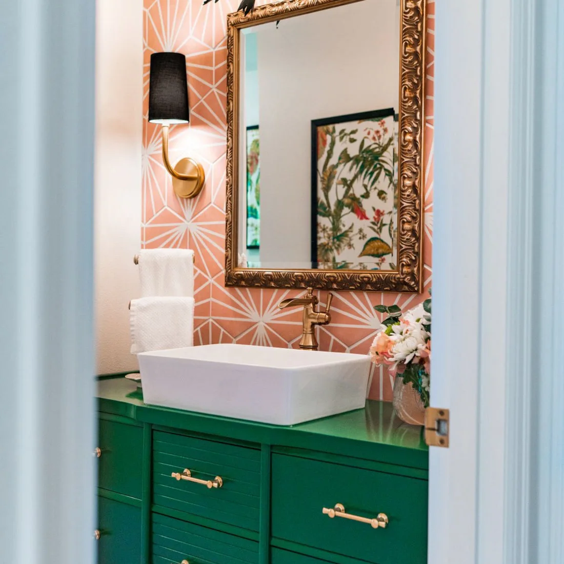 A pink geometric tile wall sits behind an ornate gold-framed mirror above a green vanity and white vessel sink