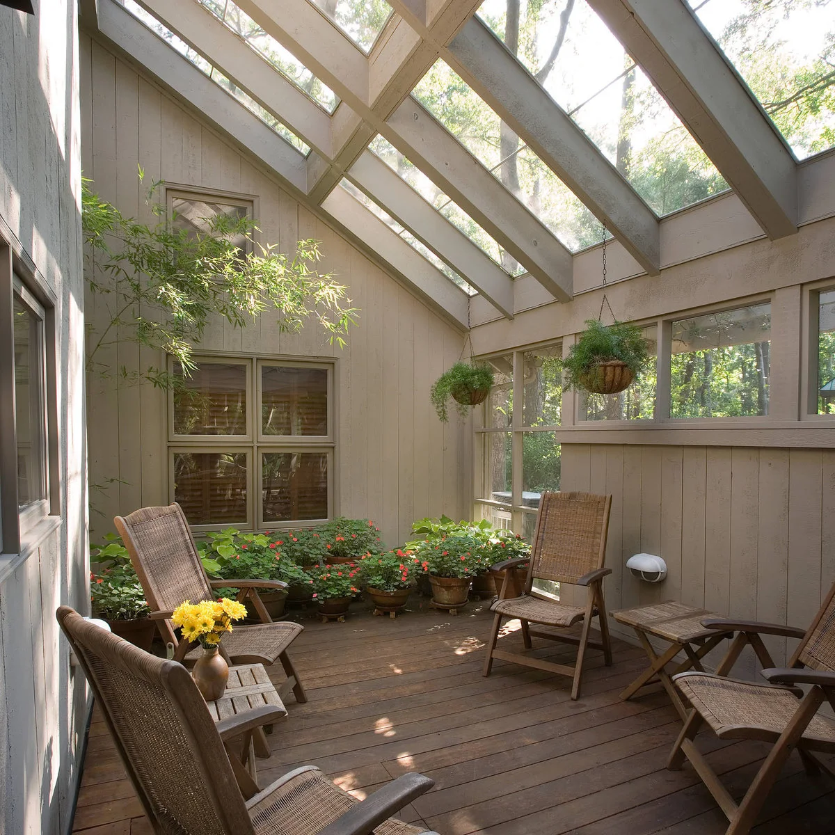 Woven chairs and small folding tables beneath a glazed roof, with hanging baskets and flowering pots along the wall.