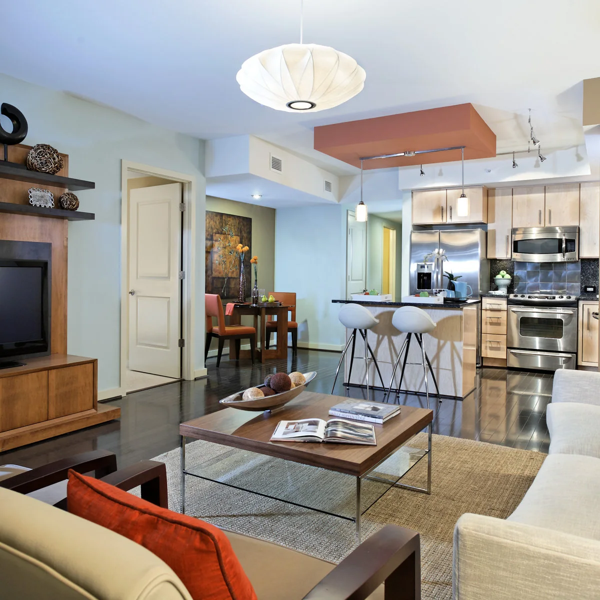 An open apartment has a pale sofa and low coffee table facing a television, with a small dining table in a side recess and white stools at the kitchen counter.
