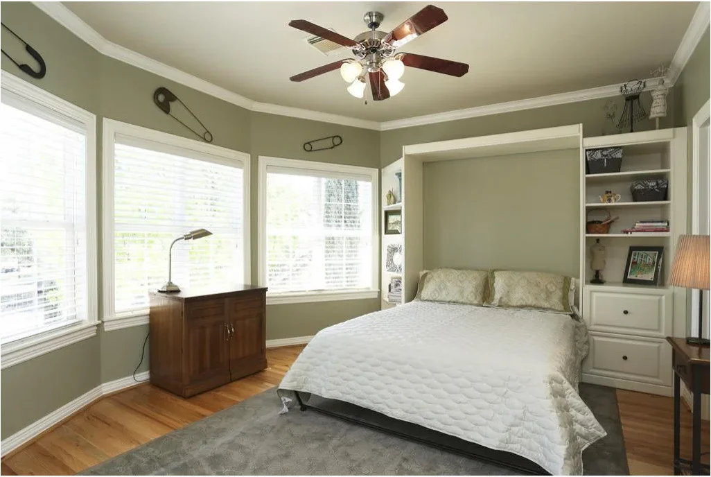 A pale wall bed is open beside white shelves and drawers, with a closed brown sewing cabinet beneath the windows.