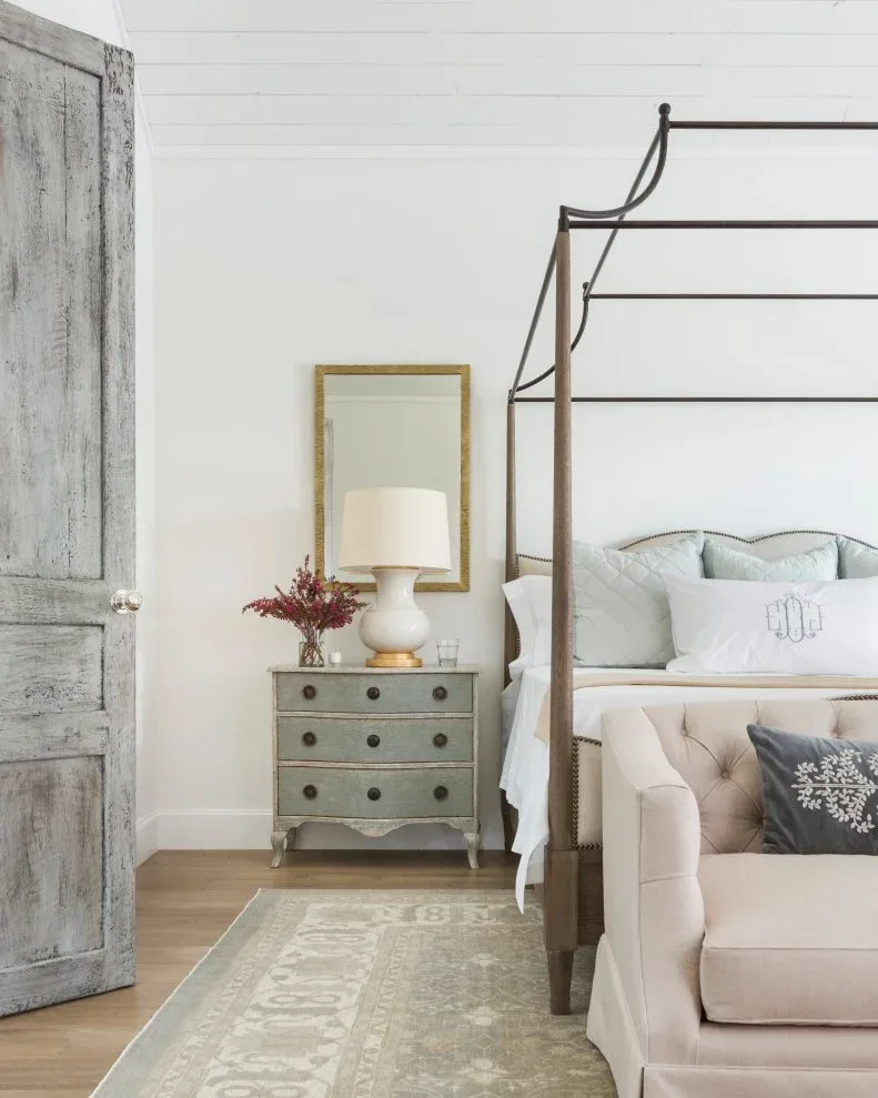 White boarded bedroom with dark canopy frame, gray bedside chest, pale pillows and warm-toned mirror
