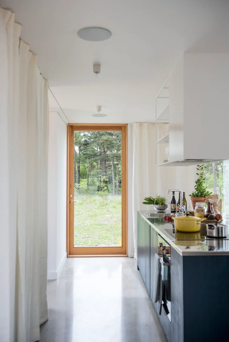 A narrow kitchen has a dark single cabinet run, white upper storage, a pale floor, a long curtain on the opposite side and a wood-framed glazed door.