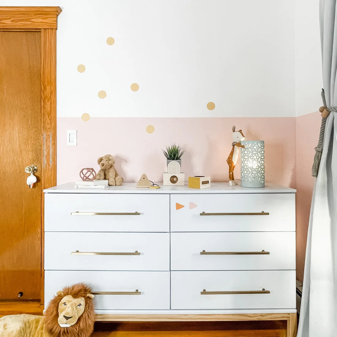 A white six-drawer chest with pale wood legs and warm-metal pulls stands against a pink lower wall with gold dots above.