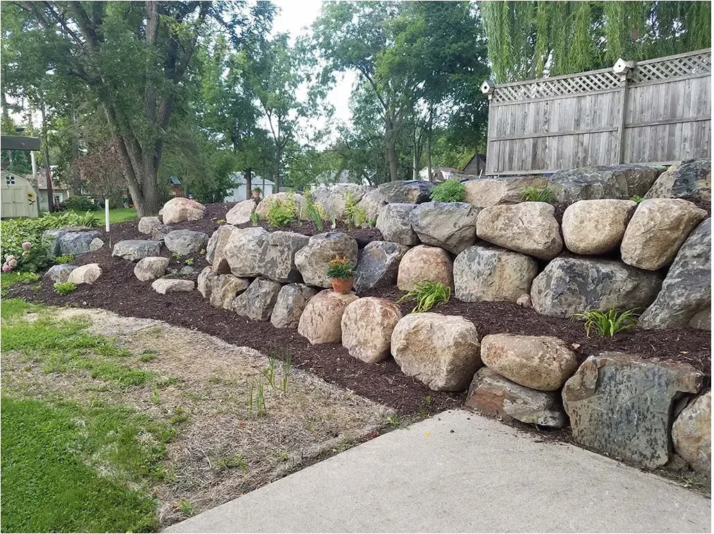 Large boulders retain a sloping mulched bed above a paved garden area and below a fence.