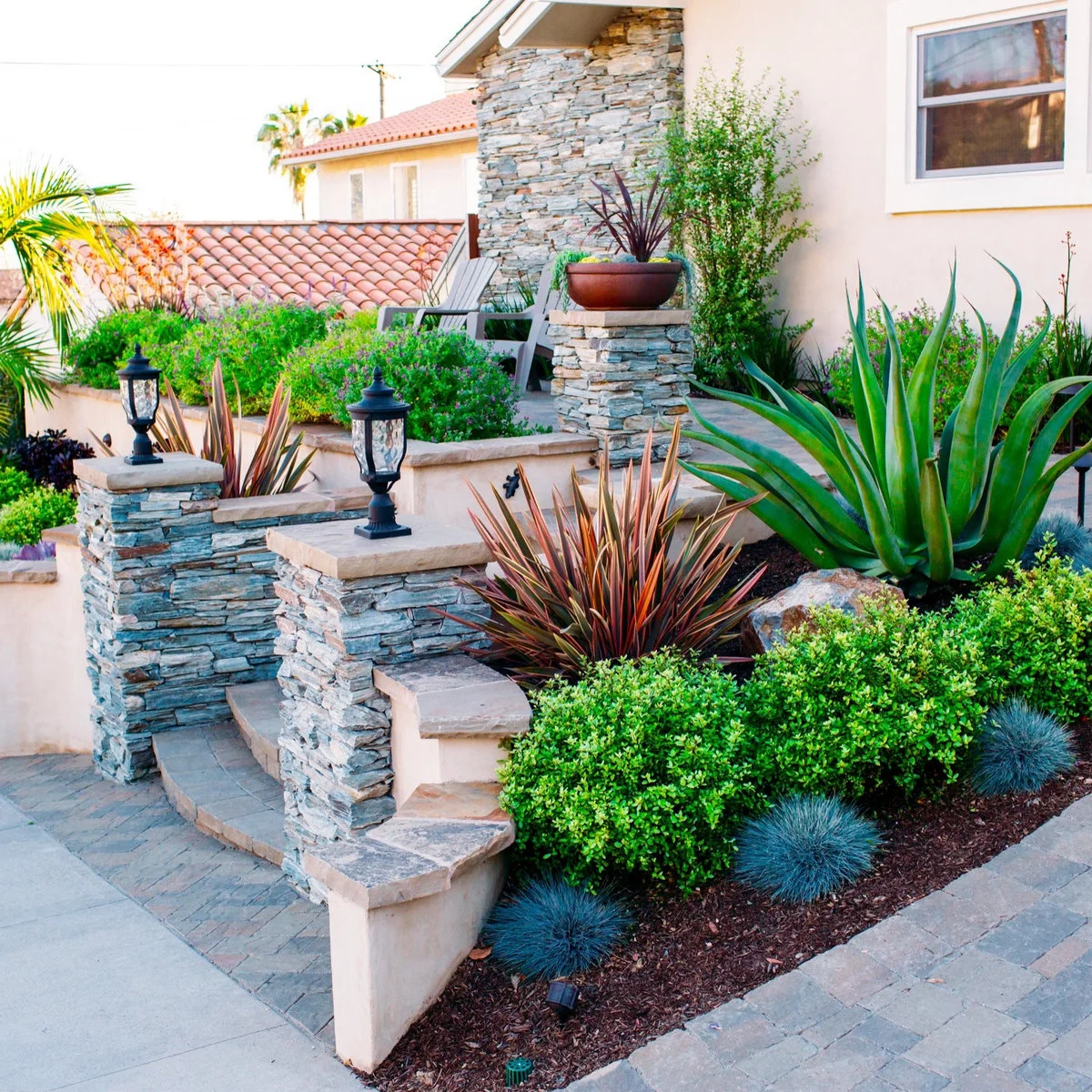 A stepped front garden with pale retaining edges, stone-faced posts, rounded shrubs and contrasting upright foliage.