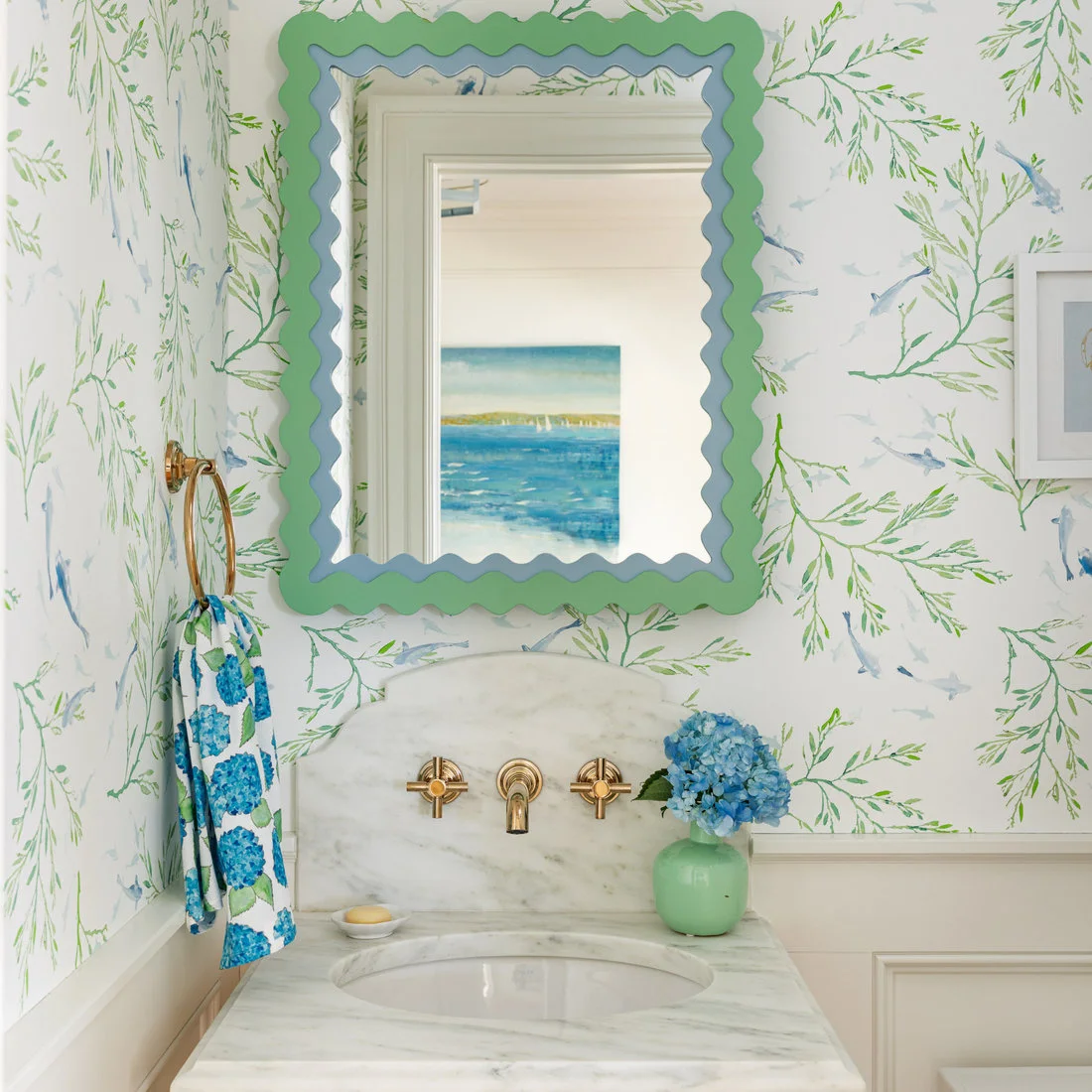 Fine blue-and-green bird wallpaper surrounds a green and blue wavy powder-room mirror above a pale sink beside a patterned towel and blue flowers