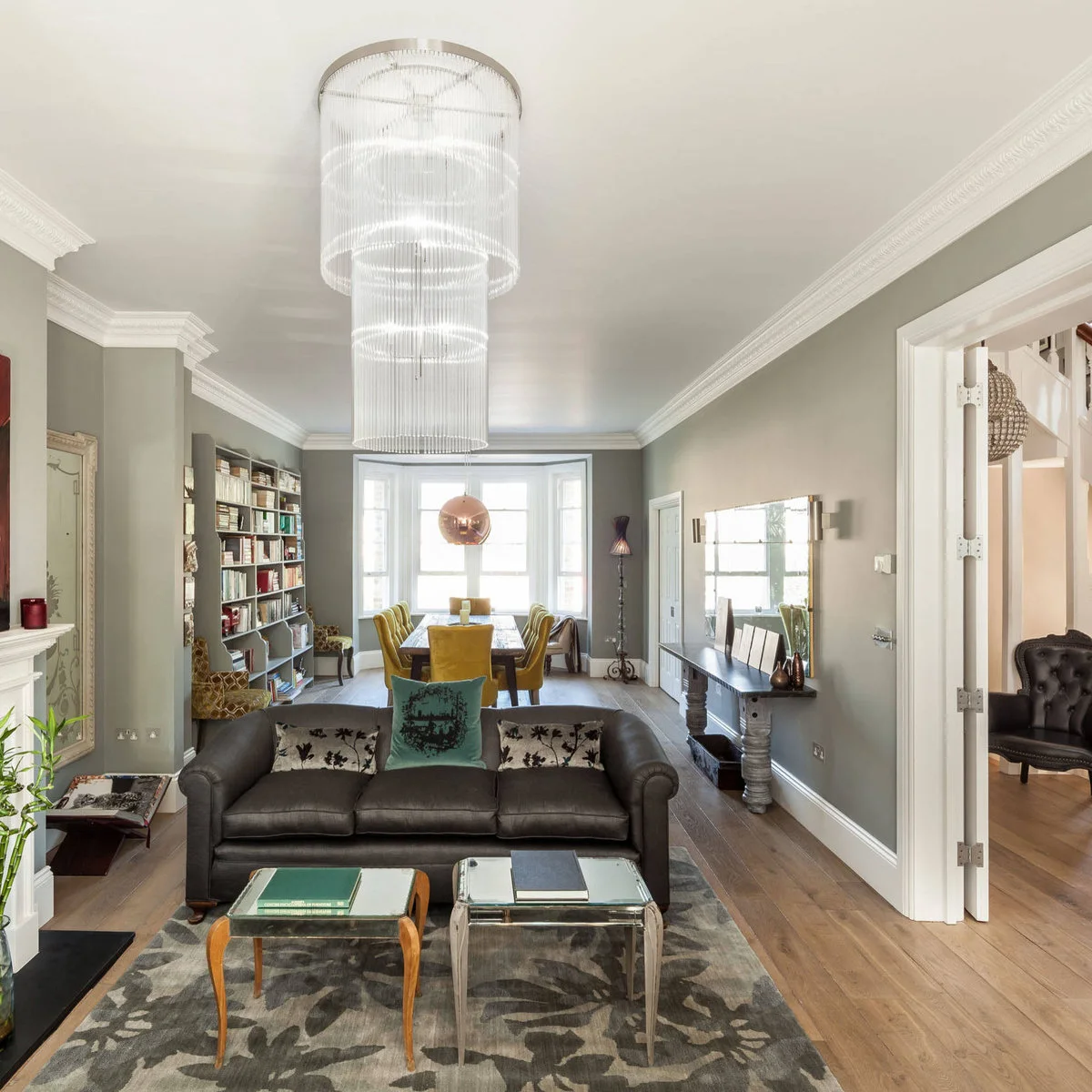 Long gray room with a dark sofa facing the foreground and a dining table with yellow chairs beside the rear bay window.