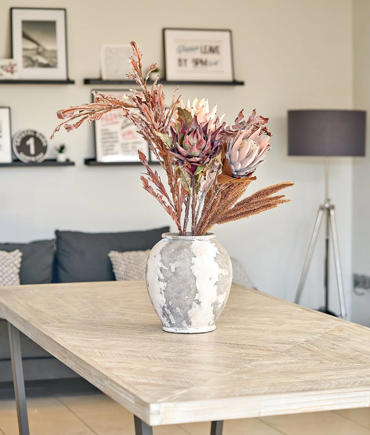 Three large dried protea heads and finer stems rise from a pale distressed vase on a timber table in a living room.