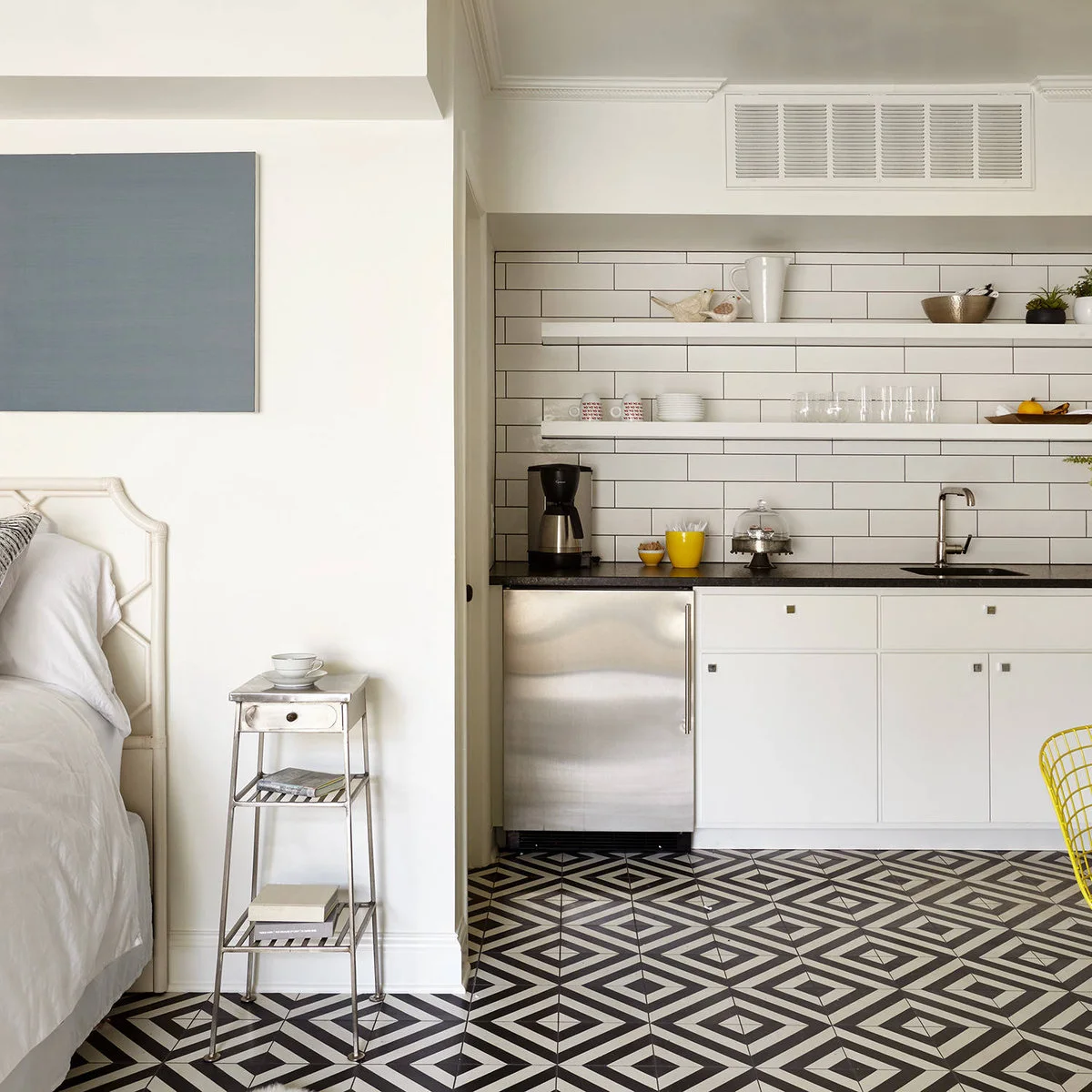 White kitchenette with an undercounter refrigerator, small sink and open shelves beside a bed