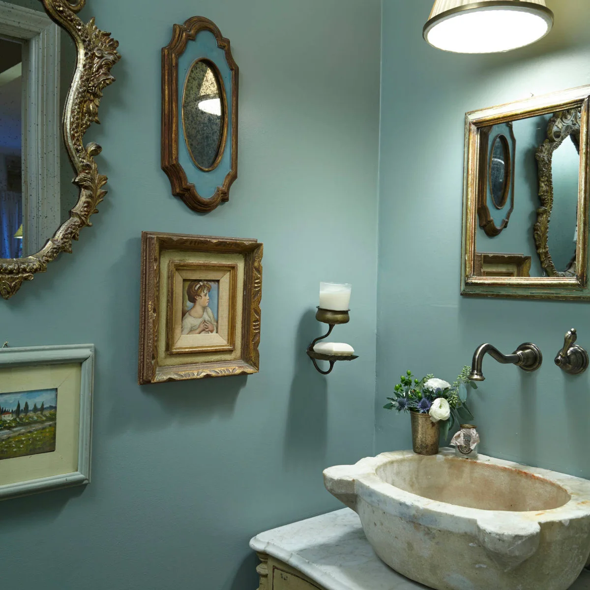 Small framed portrait and landscape among ornate mirrors on a blue-green bathroom wall