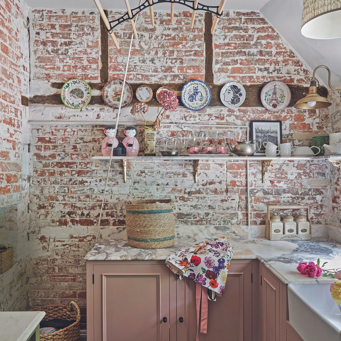Decorative plates mounted along a dark beam on a brick kitchen wall above a shelf of cups and glasses.