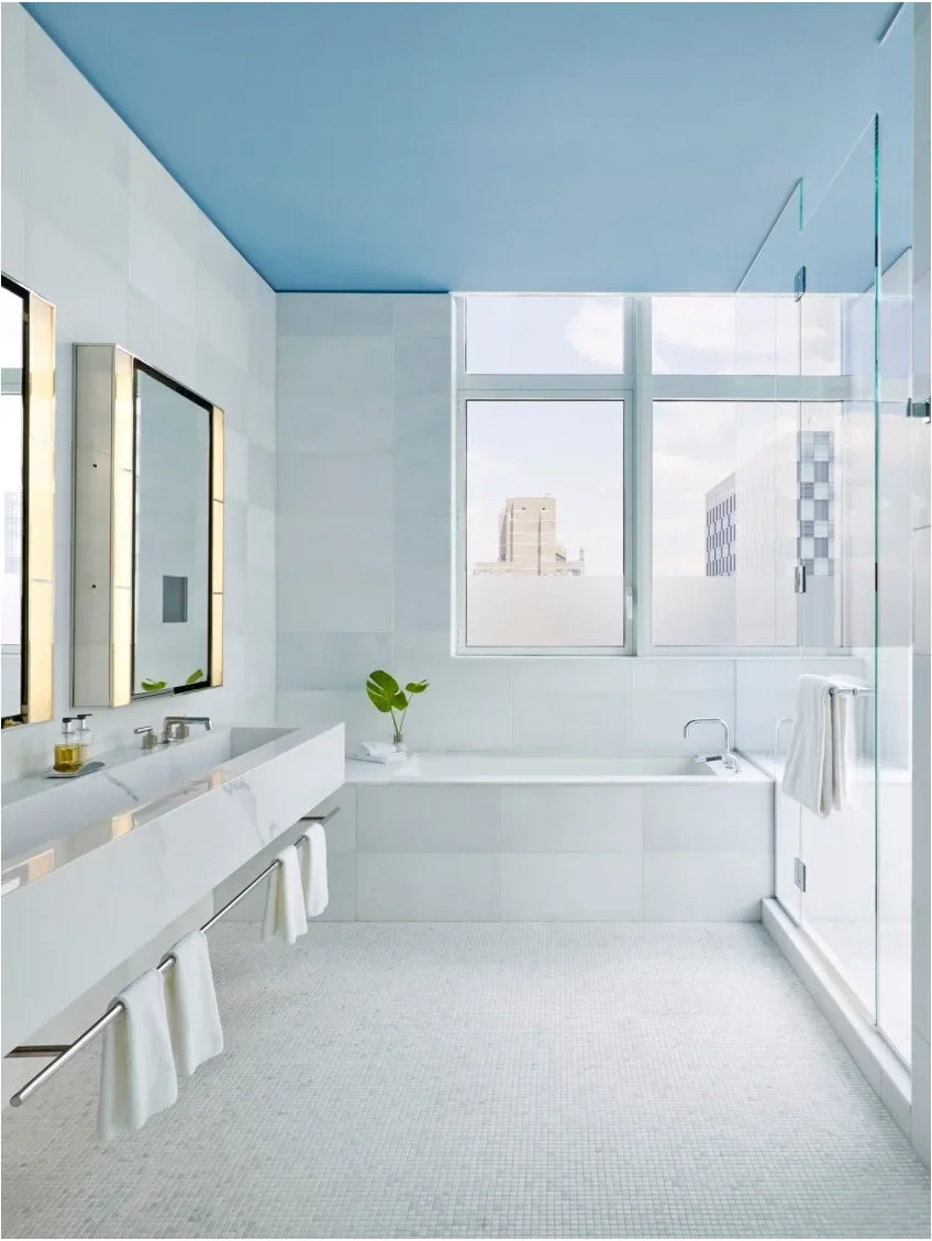 Several white towels hang from a long metal rail beneath a floating trough sink in a pale bathroom with a blue ceiling.