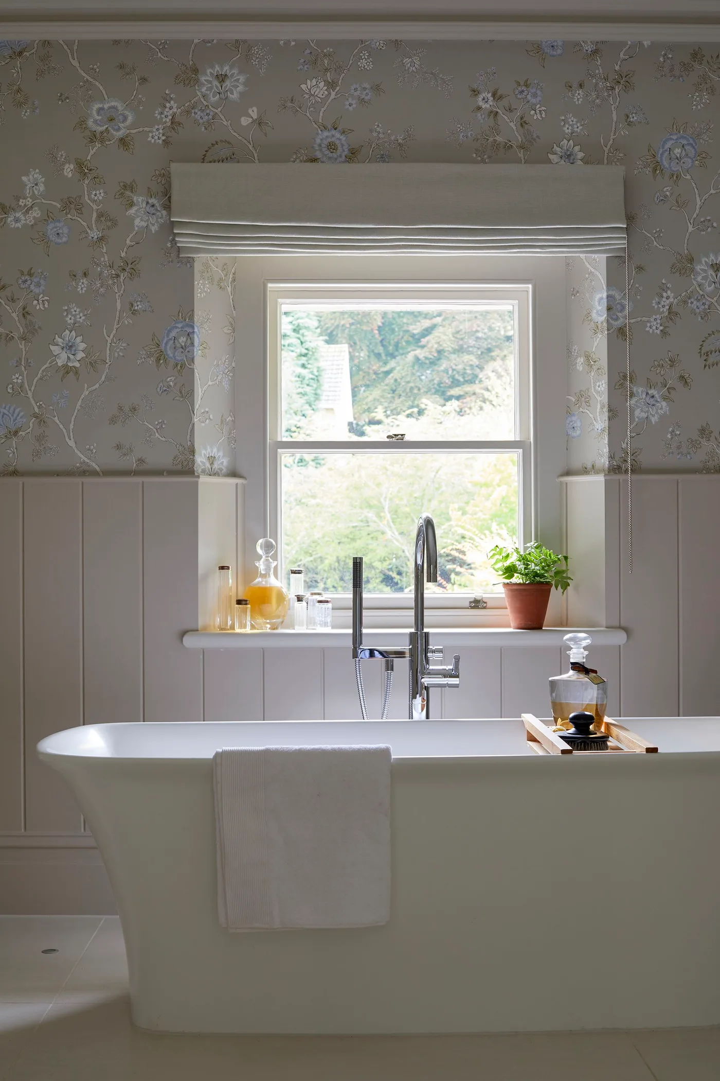Bathroom with pale wall paneling, a soft floral wall pattern, white freestanding bath and a fabric window blind.