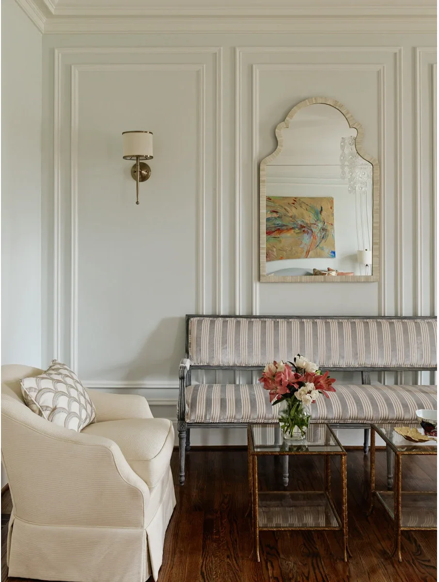 A small pale shaded wall light with a warm metal stem occupies a pale green wall panel beside a scalloped mirror and striped settee.