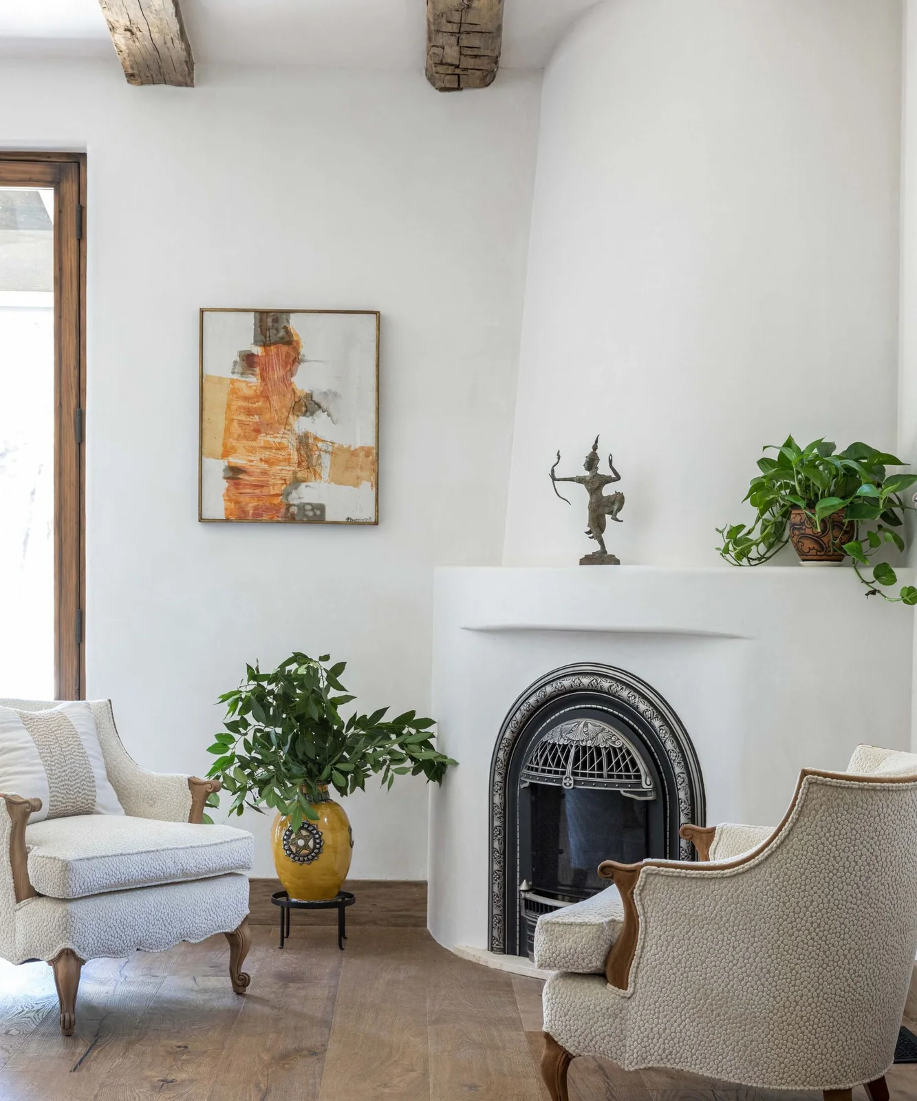 A rounded pale fireplace with a dark arched insert stands between pale curved chairs beside warm-colored artwork, greenery and exposed ceiling beams
