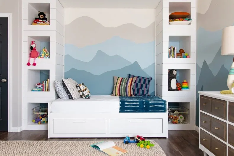 A white bench with storage drawers sits between open toy shelves against a pale blue and gray mountain mural, with patterned cushions above.