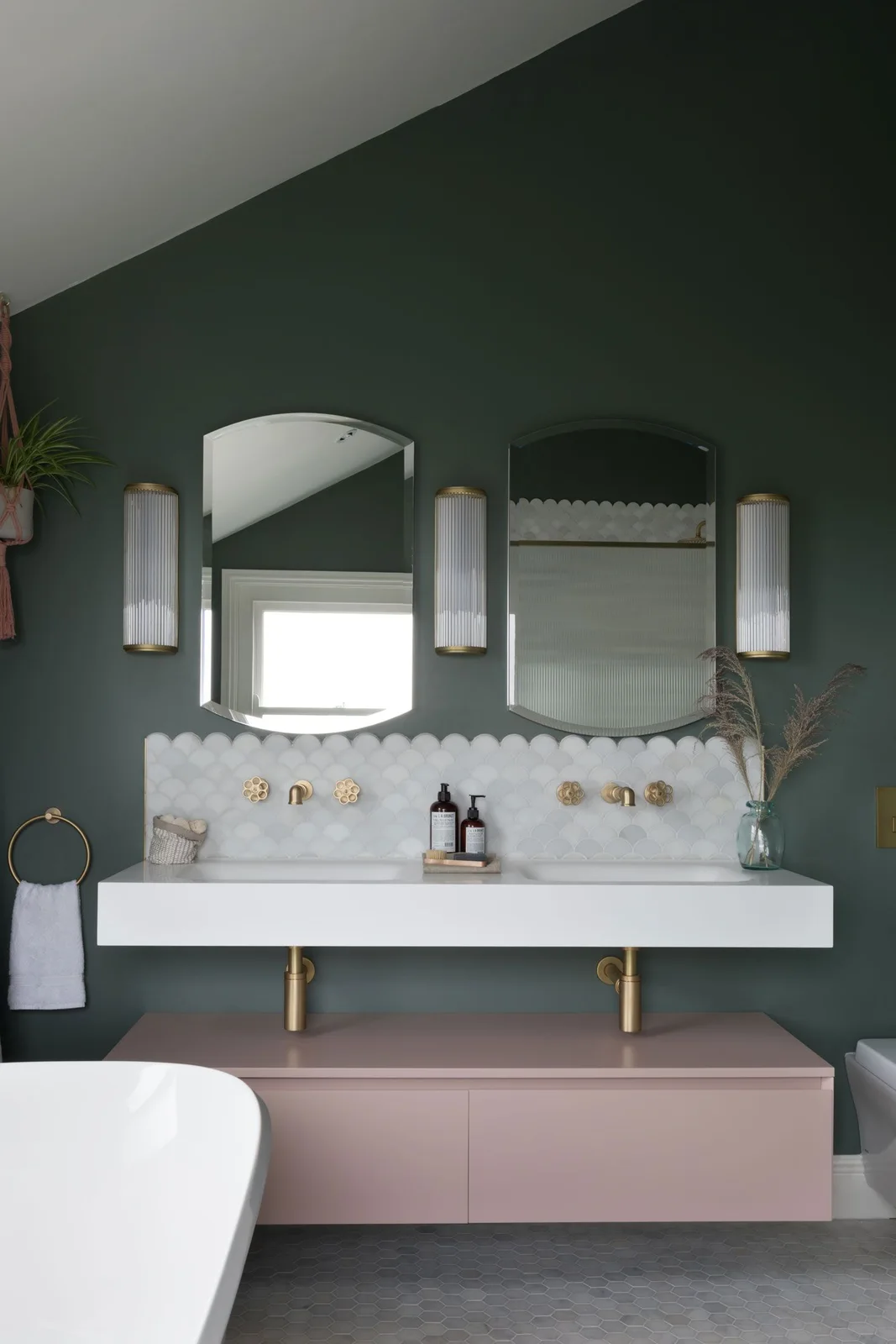 Dark green walls surround white scallop backsplash tiles and a floating pink cabinet beneath a double basin.