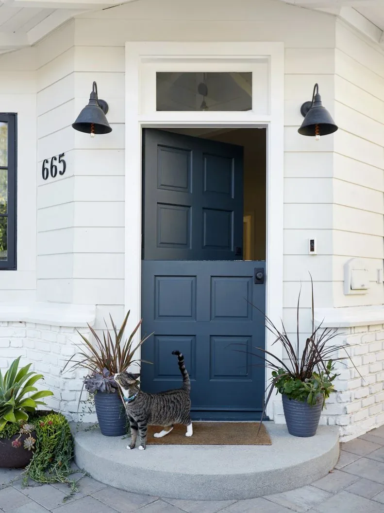 Dark blue Dutch door with white siding, paired black bell-shaped sconces, potted plants and curved landing